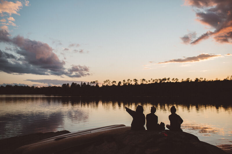 The image captures a serene sunset scene. Three silhouetted figures sit on a rock, gazing at the water. One person points towards the sky. The sky is filled with clouds, and the water reflects the colors of the sunset. The overall mood is peaceful and contemplative.